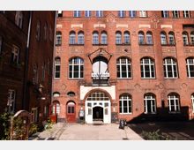 Innenhof mit mehrstöckigem Backsteingebäude und dekorativen Ziegelmustern, großen rundbogigen und rechteckigen Fenstern sowie einem zentralen Rundbogen-Eingang. Über dem Eingang hängt ein Schild „Sophiensäle“, davor liegt eine gepflasterte Fläche mit wenigen Pflanzen und Aufstellschildern.