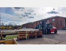 Industriegelände mit roten Backsteingebäuden, einem Gabelstapler im Vordergrund und mehreren Gütern wie Metallkisten und gelben Arbeitsfahrzeugen im Hintergrund. Auf dem Gelände sind Schienen, eine asphaltierte Fläche und vereinzelt Bäume am Rand sichtbar.