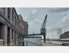 Backstein-Industriegebäude an einem Hafenbecken, mit einem alten Hafenkran im Vordergrund. Im Hintergrund sind weiße Tanks mit dem Schriftzug „Unitank“ sowie weitere Gebäude sichtbar, der Himmel ist bewölkt.