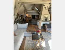 Wohnzimmer in einem Dachgeschoss mit sichtbaren Holzbalken, einer großen Ecksofa-Landschaft, Glastisch mit dekorativem Metallgestell, hellem Teppich und Kamin. Im Hintergrund befindet sich ein Essbereich mit Tisch und Stühlen sowie eine offene Küche.