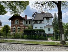 Zweistöckiges Gebäude im historischen Stil mit einer Fassade aus rotem Backstein und weiß verputzten Elementen, umgeben von einem Metallzaun und Gartenbepflanzung. Im Vordergrund sind eine gepflasterte Straße und ein großer Baum zu sehen.