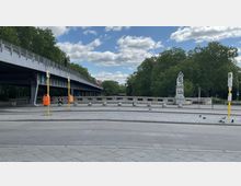 Straßenszene mit einer breiten Brücke links, unter der eine Fußgängerpassage führt. Im Vordergrund ist eine Straßenbahnhaltestelle mit gelben Pfosten und orangen Abfallbehältern. Rechts steht eine Statue auf einem steinernen Sockel, umgeben von Bäumen im Hintergrund.