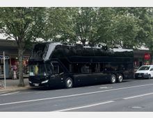Ein großer schwarzer Doppeldeckerbus parkt am Straßenrand vor einem Gebäude mit Geschäften, darunter ein Souvenirgeschäft. Im Hintergrund sind Bäume und ein Gehweg erkennbar.