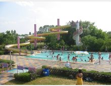 Ein Freibad mit einem großen Schwimmbecken, in dem zahlreiche Menschen baden, und einer gelben Wasserrutsche, die von pinkfarbenen Säulen gestützt wird. Im Hintergrund ist eine Wendeltreppe zu sehen, die zu der Wasserrutsche führt, und die Umgebung ist von grünen Bäumen umgeben.