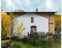 Zweistöckiges Gebäude mit heller Putzfassade in der Mitte und seitlich sichtbaren gelben Klinkersteinen, umgeben von einem Garten mit Pflanzen und einem Baum mit gelben Herbstblättern. Ein einfaches schmiedeeisernes Geländer trennt den Garten vom Gebäude.
