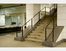 Untergrundbahnhof mit modernen weißen Fliesenwänden, einer Treppe mit Geländer in der Mitte und einer Rampe rechts. Im Hintergrund sind Gleise, eine Mülltrennstation und Betonpfeiler sichtbar.