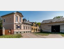 Ein kleiner gepflasterter Weg führt durch eine ländliche Szenerie mit einem rosa-bemalten, zweistöckigen Gebäude mit dekorativer Fassade auf der linken Seite und einer großen Scheune mit schwarz lackierten Doppeltüren auf der rechten Seite. Im Hintergrund sind weitere kleine Gebäude sowie Bäume erkennbar, die die ruhige Umgebung ergänzen.