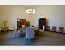 Ein schlichter Besprechungsraum mit einem großen runden Tisch in der Mitte, umgeben von gepolsterten Stühlen. Der Raum hat cremefarbene Wände, Holztüren, eine kleine Wanduhr und einen Kronleuchter an der Decke, sowie zusätzliche Stühle entlang der Wände.