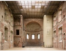 Ehemaliger Kircheninnenraum mit freigelegtem Backsteinmauerwerk, einer halbrunden Apsis im Hintergrund und modernisiertem Glasdach. Die Wände sind durch hohe, offene Fensteröffnungen geprägt, daneben befindet sich ein moderner rechteckiger Aufzugsschacht in hellem Material.