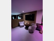 Moderner Innenraum mit lila Beleuchtung, bestehend aus zwei gepolsterten grauen Sesseln, einem runden Tisch mit weihnachtlichem Adventskranz in der Mitte, und einer Wand mit Holzpaneelen sowie einem flachen Bildschirm. Im Hintergrund ein Kamin und eine dekorative Pflanze.