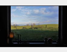 Blick aus einem Fenster auf eine weitläufige grüne Wiese mit wenigen Bäumen und einem Zaun im Hintergrund unter einem blauen Himmel mit vereinzelten Wolken. Auf der Fensterbank stehen eine Lampe mit Schirm, Kerzenhalter und Dekorationsobjekte.