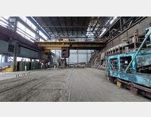 Innenraum einer großen Industriehalle mit hohen Decken und Stahlträgern. Im Vordergrund befinden sich Gleise und ein alter, blauer Industriewagen, während die Halle von Maschinen, Rohren und einer gelben Krananlage dominiert wird.