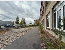 Seitenansicht eines länglichen Industrie- oder Lagergebäudes mit verputzter Fassade und großen Fenstern entlang eines schmalen, rissigen Gehwegs. Links liegt ein eingezäunter Parkplatz und eine überwachsene, teilweise gepflasterte Fläche; im Hintergrund stehen Bäume unter bewölktem Himmel.