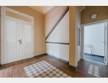 Innenflur eines Altbaus mit hohen Decken, hellen Wänden und dunklem Holzboden mit gemusterten Fliesenläufern. Links eine große weiße Doppeltür, rechts ein Durchgang zu einem hellen Raum; an der Wand ein weißer Treppenunterbau mit dunkler Zierleiste.