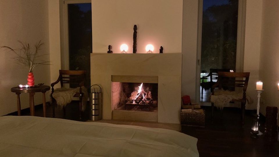 Gemütliches Schlafzimmer mit großem Bett im Vordergrund und offenem Kamin in einer schlichten, hellen Wandnische. Links und rechts stehen Sessel und kleine Tische mit Kerzen; hohe Fenster zeigen die dunkle Außenansicht.