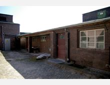 Backsteingebäude in einem Hof mit Kopfsteinpflaster, erkennbar an Fensterfronten, einer roten Holztür und einem Vordach mit Regenrinne. Im Hintergrund weitere Gebäudeteile aus Backstein mit einer Metalltür.