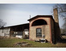 Backstein-Industriegebäude mit großem Rundbogen im Obergeschoss und hohem Schornstein, daneben eine offene überdachte Halle mit Stützen. Davor liegt eine verwilderte Rasenfläche mit Laub und vereinzelten Gegenständen; im Hintergrund sind kahle Bäume zu sehen.