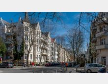 Straßenszene in einem Wohngebiet mit mehrstöckigen Altbauhäusern und einer gepflasterten Straße. Barock-inspirierte Fassaden mit Balkons säumen die Straße, während kahle Bäume und einige geparkte Autos den Gehweg begrenzen.