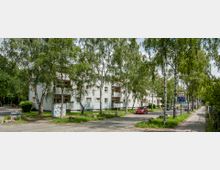 Wohnstraße mit mehrstöckigen, hellen Wohngebäuden im Hintergrund, umgeben von hohen Bäumen. Gehwege, geparkte Autos und ein aufgestelltes Verkehrsschild prägen die Umgebung.