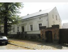 Ein historisches zweistöckiges Wohngebäude mit heller Putzfassade, verzierten Fensterrahmen und detailliertem Stuckdekor. Im Vordergrund sind ein schmiedeeiserner Zaun, ein gemauerter Torbogen aus rotem Backstein mit Kreuzornamenten sowie ein gepflasterter Gehweg zu sehen.