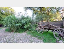 Ein hölzerner, altertümlicher Wagen mit großen Speichenrädern steht auf einer Wiese neben dichtem hohen Gras und Bambus. Im Hintergrund sind Bäume und ein gepflasterter Weg sichtbar.