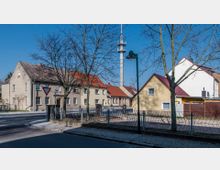 Straßenszene in einem Wohnviertel mit mehreren Häusern in hellem Putz und roten Dächern, im Hintergrund ist ein hoher Funkturm sichtbar. Bäume ohne Laub und ein Zaun begrenzen den Gehweg im Vordergrund.
