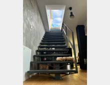 Stählerne Innentreppe mit modernen, offenen Stufen, die zu einem Dachfenster führt, durch das blauer Himmel und Wolken sichtbar sind. Die Umgebung ist ein minimalistisch gestalteter Raum mit Holzboden und grauer Wand.