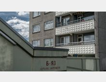 Wohnblock aus Betonplattenbauweise mit weißen Balkonen, die ein geometrisches Muster zeigen, im Hintergrund helle Fensterreihen; im Vordergrund ein graues Gebäude mit kyrillischer Beschriftung.