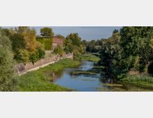 Backsteinbau mit teilweise bewachsener Fassade am Ufer eines Flusses, umgeben von dichter grüner Vegetation und Bäumen; der Fluss ist gesäumt von einer Betonmauer auf einer Seite und einem natürlichen Ufer auf der anderen.
