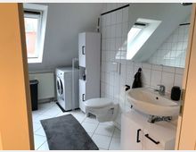 Modernes Badezimmer mit weiß gefliesten Wänden und schräger Decke mit Dachfenstern. Im Raum befinden sich ein Waschbecken mit Unterschrank, eine Toilette, eine Waschmaschine und ein Heizkörper; der Boden ist mit weißen Fliesen und einem grauen Teppich ausgestattet.