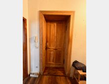 Eine massive Holztür mit natürlicher Holzmaserung in einem Türrahmen aus passendem Holz, in einem Eingangsbereich mit weiß verputzten Wänden und Holzboden. Links neben der Tür befindet sich eine Gegensprechanlage.