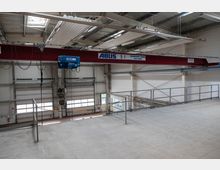 Eine industrielle Halle mit einer roten Krananlage der Marke "ABUS" (5 Tonnen Traglast) an der Decke. Im Hintergrund sind mehrere Rolltore, Geländer und eine Betonplattform zu sehen.