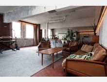 Großer, loftartiger Wohnraum mit rohen Betonwänden, sichtbaren Lüftungsrohren und großen Fenstern mit Vorhängen. Im Vordergrund stehen braune Ledersofas und ein Holztisch auf einem Teppich; im Hintergrund sind Pflanzen, Regale und Musikinstrumente zu sehen.