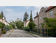 Ruhige Wohnstraße mit Einfamilienhäusern im Stil der frühen 20. Jahrhunderts, flankiert von kleinen Vorgärten und Gehwegen. Die Straße ist asphaltiert und von Bäumen sowie vereinzelten Laternen gesäumt.