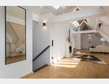 Offener Wohnbereich in einem Dachgeschoss mit weiß gestrichenen Wänden, sichtbaren Dachbalken und hellem Holzfußboden. Der Raum umfasst ein modernes Sofa, einen Kamin, eine Dekoration aus Holz sowie einen Teppich aus Kuhfell.