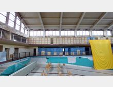 Ein leerstehendes, ehemaliges Hallenbad mit einer leeren Schwimmbeckenmulde, umgeben von verblassten türkisfarbenen Fliesen. Im Hintergrund sind Umkleidekabinen und große Fenster mit Rostspuren an den Wänden, sowie ein gelber Stoffvorhang rechts sichtbar.