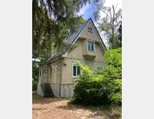 Zweistöckiges Holzhaus mit Satteldach und heller Fassade, umgeben von Bäumen und Sträuchern. Eine kleine Veranda mit Treppe führt zum Eingang, und die Umgebung ist naturbelassen mit gepflastertem Boden.