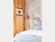 Schlafzimmer mit pastellfarbenem Dekor, einer Holzgittertrennwand mit Pflanzentopf, einer Wand mit Flamingo-Muster und einem Bett mit blauer Bettwäsche und weißer Tagesdecke im Vordergrund.