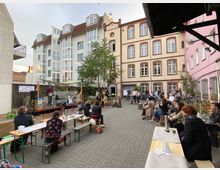 Ein Hof mit Pflastersteinen, umgeben von historischen Gebäuden mit großen Fenstern, dient als Veranstaltungsort. Im Vordergrund sitzen Menschen auf Bänken und Tischen, während auf einer kleinen Bühne ein Musiker spielt; im Hintergrund stehen weitere Zuschauer.