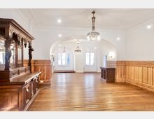 Heller, leerer Saal mit Holzdielenboden und umlaufender Holzvertäfelung an den Wänden, verbunden durch breite Rundbögen zu einem hinteren Raum mit Fenstern. An der Decke hängen mehrere Kronleuchter, links steht ein großes, reich geschnitztes Holzbuffet.