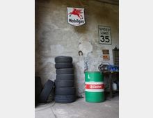 Ein Werkstattraum mit abgenutzter Wand zeigt gestapelte Autoreifen, eine grüne Öltonne mit "Castrol"-Logo, ein altes „Mobilgas“-Schild an der Wand sowie ein Geschwindigkeitsschild mit der Aufschrift „Speed Limit 35“. Im Hintergrund sind weitere Werkstattutensilien wie Kabel und Plakate sichtbar.