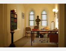 Ein kleiner Raum mit gewölbter Decke und drei verzierten, farbigen Glasfenstern im gotischen Stil. Im Vordergrund ein Schreibtisch mit Bürostuhl sowie Bücherregale und eine Büste auf einem Sockel im Hintergrund.
