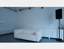 Ein minimalistisch eingerichteter Raum mit weiß gestrichenen Wänden, einer weißen Ledercouch im Vordergrund, einem schwarzen Lautsprecher auf einem Stativ rechts und weißem Mobiliar im Hintergrund. Die Atmosphäre wirkt schlicht und funktional.