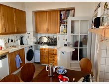 Kleine Küche mit weißen Fliesen und Schränken aus Holz. Zu sehen sind unter anderem eine Kaffeemaschine, ein Kühlschrank, eine Waschmaschine, ein Esstisch mit Geschirr und Tomaten sowie eine Glastür mit Sprossenrahmen.