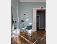 Ein heller Raum mit hellblauen Wänden, einer weißen Doppeltür und einem Holzboden in Fischgrätmuster. Im Vordergrund stehen ein moderner drehbarer Stuhl, ein kleiner runder Beistelltisch mit Dekoration und eine große angelehnte Spiegelglasscheibe.