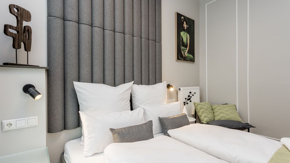 Modernes Schlafzimmer mit zwei zusammengestellten Einzelbetten, weißer Bettwäsche und einem hohen, gepolsterten Kopfteil aus grauen Stoffpaneelen. Helle Wände mit Zierleisten, Wandlampen, ein gerahmtes Bild sowie eine kleine Sitzbank mit grünen Kissen ergänzen den Raum.