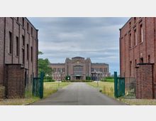 Backstein-Gebäudekomplex mit einer symmetrischen, breiten Hauptfassade im Hintergrund, flankiert von zwei schmalen Nebengebäuden im Vordergrund. Eine asphaltierte Zufahrt führt durch ein offenes Tor zur zentralen Eingangstreppe des Hauptgebäudes.