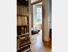 Blick durch eine geöffnete Tür in ein helles Zimmer mit einem großen Fenster, durch das viel Tageslicht einfällt. Im Vordergrund steht ein Bücherregal, und im Hintergrund ist ein Sitzsack, eine Stehlampe und ein Holzboden mit Fischgrätmuster zu sehen.