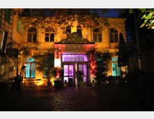 Beleuchtetes historisches Gebäude mit zwei Stockwerken in gelber Fassade, großen Fenstern und einem mittig gelegenen Eingang. Der Vorderhof ist mit Kopfsteinpflaster bedeckt, umgeben von Pflanzen, und farbige Lichter (rot, violett, blau) akzentuieren die Architektur.
