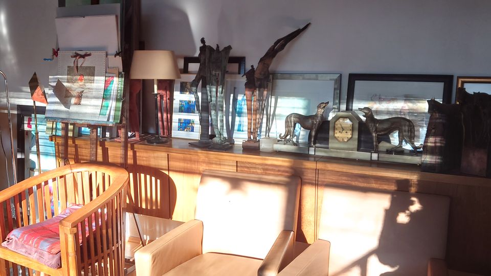 Ein gemütlicher Innenraum mit Holzmöbeln, darunter zwei beige Polstersessel und ein Holzstuhl mit buntem Kissen. An der Wand stehen dekorative Skulpturen, Bilderrahmen und gemalte Leinwände, beleuchtet von einfallendem Sonnenlicht.